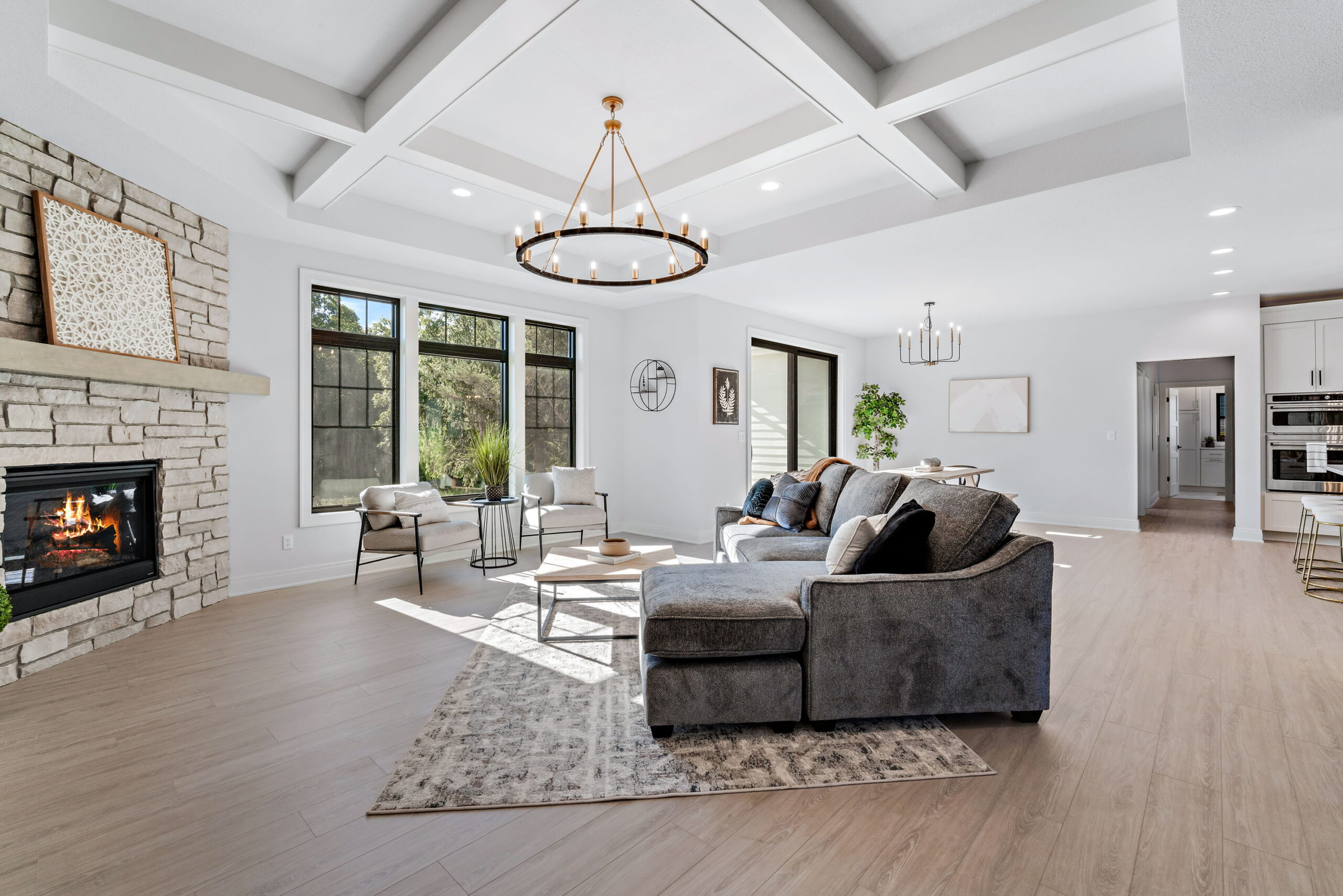 Bright living room with stone fireplace, coffered ceiling, large windows, and open floor plan